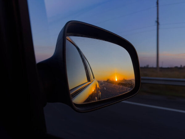 Sunset reflection in a car side mirror while driving on a highway, capturing warm evening light, road lines, and a peaceful travel atmosphere.