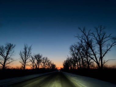 Alacakaranlıkta kış yolu dramatik mavi ve turuncu eğimli gökyüzü hareket bulanık ağaçları ve boş kırsal yol. Atmosferik alacakaranlık manzara arkaplanı.