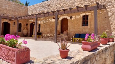 Geleneksel Akdeniz taş evinin güneşli avlusunda çiçek açan tencereler, kırsal mimari, ahşap pergola ve parlak gün ışığında yakalanan canlı yaz atmosferi.