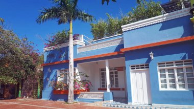 Beyaz desenli, turuncu aksanlı, tropik bitkiler ve bir palmiye ağacıyla çerçevelenmiş, parlak mavi gökyüzü altında güneşli bir sokak sahnesinde büyüleyici bir mavi koloni tarzı ev.