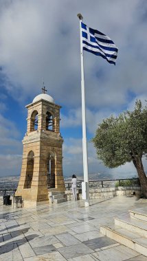 Yunanistan 'ın başkenti Atina' daki Lycabettus Tepesi 'nin üzerinde parçalı bulutlu bir gökyüzü altında sallanan beyaz kubbeli taş çan kulesi ve panoramik şehir manzarasına hayran tek bir ziyaretçi.