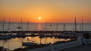 Gün batımında tekneler ve yelkenlilerle dolu bir marina manzarası. Güneş sakin bir denizin ve parlayan turuncu bir gökyüzünün üzerinde batıyor. Huzurlu deniz manzarası seyahat ve yaşam tarzı temaları için mükemmel.