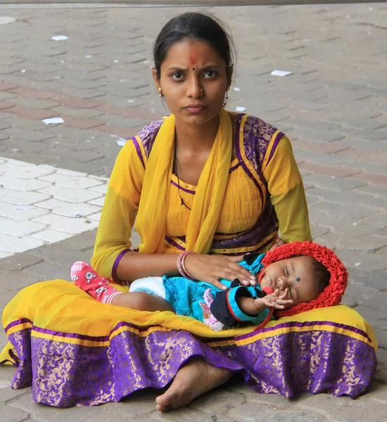 Bangalore, Hindistan - 01 Kasım 2017: Geleneksel giyimli Hintli bir kadın kaldırımda çıplak ayakla oturuyor, kucağında uyuyan bir bebek tutuyor. Sahne, sessiz bir esneklik ve anne bakımı anı yakalıyor.