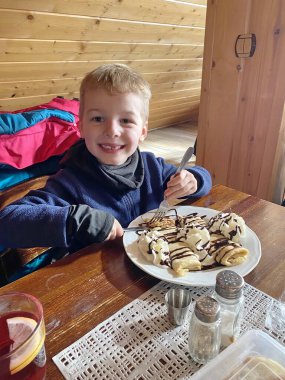 Sıcak bir kafede ya da pansiyonda, ahşap bir masada krema ve çikolata soslu tatlı kreplerin tadını çıkaran mutlu çocuk. Çocuk bir çatal tutuyor ve kameraya bakıyor, sıcak ve neşeli bir atmosfer yaratıyor. Saf ahşap iç döşeme...