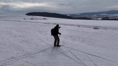 Karlı kış manzarasında karla kaplı açık bir arazide kayak yapan yalnız bir insan. Yuvarlanan tepeler ve bulutlu bir gökyüzünün altındaki uzak dağlar. Büyük bir sırt çantası taşıyan ve uzun adımlarla ilerlerken kayak sopaları kullanan kişi..