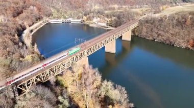 Sonbahar ormanlık ve kırsal kesimlerle çevrili sakin bir nehir üzerinden çelik bir demiryolu köprüsünü geçen bir yolcu uçağının görüntüsü, yüksek açılı bir perspektiften çekildi..
