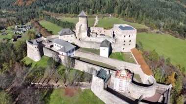 Yoğun sonbahar ormanı ve dağ manzarasıyla çevrili, yüksek açılı drone perspektifinden çekilmiş, iyi korunmuş bir ortaçağ taş şatosunun havadan görünüşü..