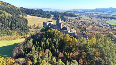 Ortaçağ taşından bir şatonun insansız hava aracı görüntüsü. Stara Lubovna ormanlık bir tepeye tünemiş, açık mavi gökyüzünün altında sonbahar yapraklarıyla çevrili, tarihi, mirası ve sakin doğa manzarasını aktarıyor..