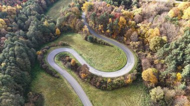 Yamaçtan geçen dolambaçlı bir yolun havadan görünüşü. Yol, ön planda çarpıcı bir viraj alıyor ve üzerinde bir araba gidiyor. Yolu çevreleyen yoğun sonbahar renkleri, bir sürü kırmızı, portakal, sarı ve