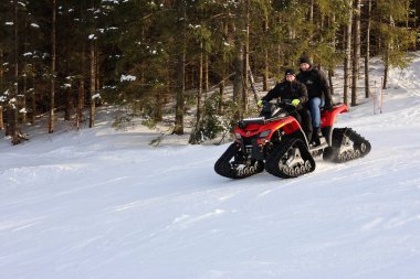 İki adam, kışın ortasında yeşil ağaçlarla çevrili, karlı bir orman patikasında kırmızı izli bir ATV 'ye biniyor. Soğuk iklimde açık hava maceralarını ve off-road ulaşımını yakalıyorlar..