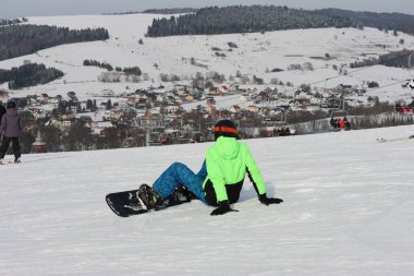 Parlak yeşil ceketli snowboardcu, karlı bir yamaçta dinleniyor, tepeye bakan bir köyde ve koltukta oturuyor, geniş açılı, kış sporunu, dağ yaşam tarzını ve kırsal manzarayı yakalıyor..