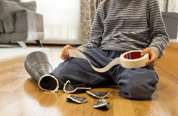 Elinde bir rulo yapışkan bant olan yaramaz bir çocuk bir topla kırdığı vazoyu tamir etmeye çalışıyor.