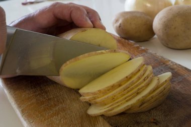 Koyu ahşap bir yüzey üzerinde keskin çelik bir bıçakla taze bir patatesi ince ince dilimleyen birinin ayrıntılı görüntüsü..