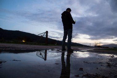 Yağmur birikintisinin yanında duran bir adamın silüeti aydınlanmış cep telefonu ekranına bulutlu bir alacakaranlık gökyüzüne bakıyor..