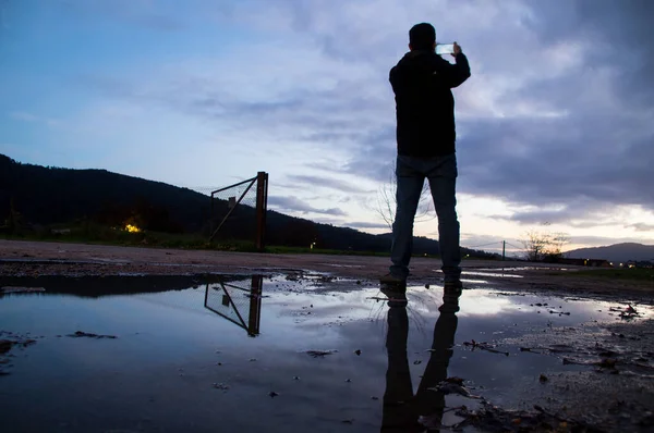 Kırsal bir alanda duran bir adam, telefonuyla yağmur suyu birikintisine yansıyan ufkun resmini çekiyor..