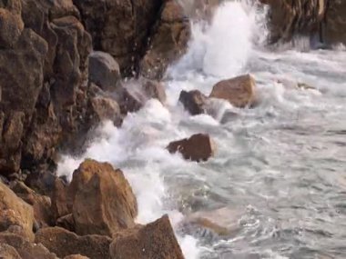 Yumuşak gün ışığında dalgaların kayalara vurduğu engebeli bir sahil manzarası..