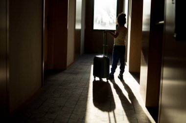 Bagajlı çocuk güneşli otel koridorunda duruyor.