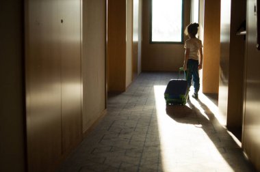 Bir çocuğun bavul sapını tutarken yandan görünüşü, dramatik yan aydınlatma ve halı kaplı zeminde uzun gölgeler ile yakalanmış..