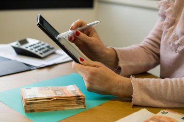 Elinde tablet ve kalemle pembe bluzlu bir kadın ön planda 50 Euro 'luk banknotlarla hesapları inceliyor..