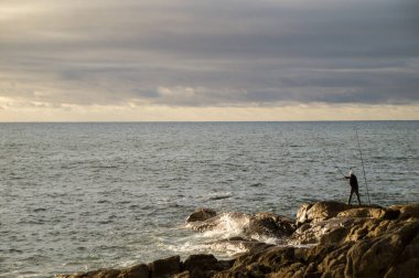 Gün batımında dramatik bulutlu bir gökyüzünün altında büyük deniz kayalarında balık tutan bir adamın geniş açılı görüntüsü..