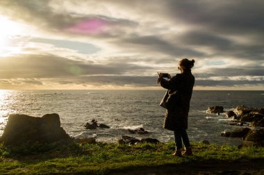 Çimenli bir uçurumun kenarında duran bir kadın akıllı telefonuyla Atlantik Okyanusu 'ndaki altın saati yakalıyor.