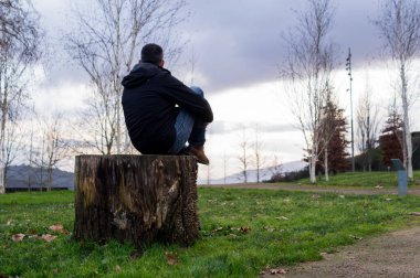 Parktaki büyük bir ağaç kütüğünde tek başına oturan bir adamın dikiz aynasından gün batımında uzak dağlara bakan ve yansıtan görüntüsü..