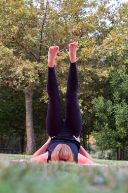 Siyah taytlı bir kadın açık havada pembe bir paspasın üzerinde Sarvangasana ya da omuz duruşu yapıyor. Çekirdek kuvveti ve dönüşümüne odaklanıyor..