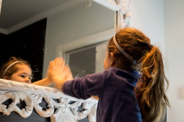 Kahverengi at kuyruğu ve saç bandı takmış genç bir kızın, tuvaletteki büyük, süslü beyaz bir aynadaki yansımasına bakışıyla..