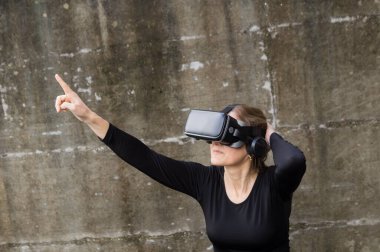 VR kulaklık takan ve parmağını gökyüzüne doğru gösteren bir kadın görünmez bir dijital arayüzle etkileşime giriyor..