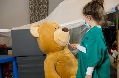Yeşil tıbbi kıyafetli bir çocuk elinde sehpanın yanındaki tıbbi aletle dev bir oyuncak ayının önünde duruyor.