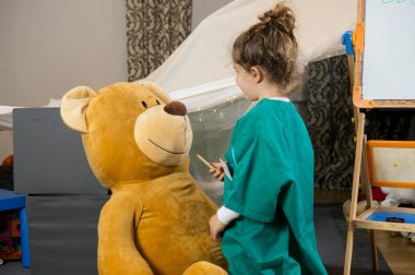 Yeşil önlük giymiş, yerde diz çökmüş ve oyun odasındaki beyaz tahtanın yanında oyuncak ayıyı muayene eden bir çocuğun tam pozu.