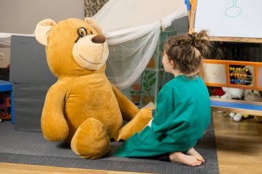 Dev oyuncak ayı ve eğitim tahtasıyla doktorculuk oynayan bir çocuk.