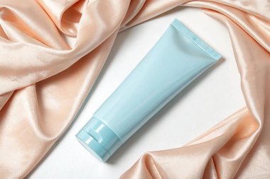 Açık mavi bir kozmetik tüp, beyaz bir yüzeyde örtülmüş şeftali rengi ipek kumaşla çevrelenmiştir. Yukarıdan çekilen, yumuşak ışıklandırma.