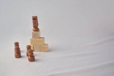 Kahverengi tahta bir satranç şövalyesi, önderliği ve stratejik hiyerarşi kavramını sembolize eden, beyaz arka planda üç piyon bulunan, hafif tahta bloklardan oluşan bir piramidin tepesinde duruyor..