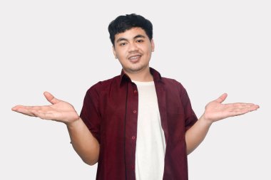 Genç yetişkin bir erkek, beyaz bir tişörtün üzerine kahverengi bir gömlek giyiyor, iki eli açık gülümsüyor ve el kol hareketi yapıyor, avuç içleri açık, açık gri bir arkaplanda izole edilmiş,.