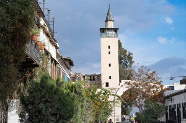 Damascus, Syria. December 10th 2025. A tall, square stone minaret with a pointed cap rises above the historic rooftops and autumn trees of old Damascus.