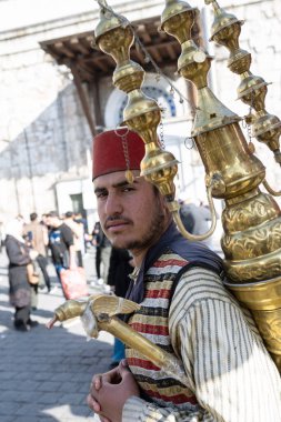 Şam, Suriye. 4 Aralık 2025. Kırmızı fes takan ve geleneksel çizgili yelek giyen bir adam sırtında büyük, süslü bir pirinç suyu makinesi taşır..