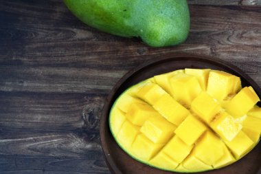 Dilimlenmiş sarı mango yarısı yeşil bir mangonun yanında küçük bir ahşap tabakta oturur..