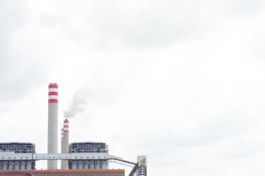 Elektrik hatları ve bacaları olan sanayi kompleksi bulutlu bir gökyüzü altında duman yayıyor.