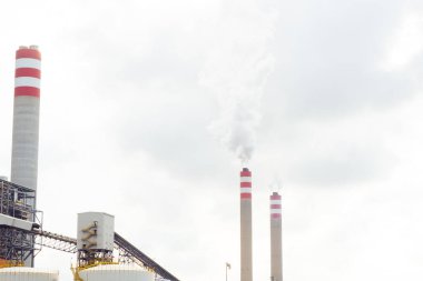 Elektrik hatları ve bacaları olan sanayi kompleksi bulutlu bir gökyüzü altında duman yayıyor.