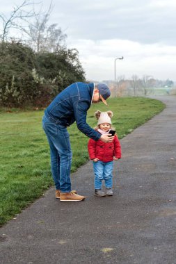 Father showing something on a smartphone to a small child on a paved path in a park.