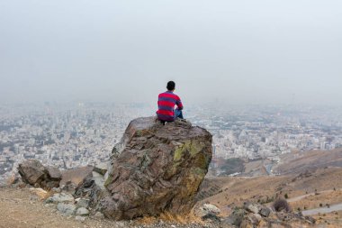 Sonbaharda Tochal dağında büyük bir kayanın üzerinde oturan genç bir turist İran 'ın Tahran şehrine bakıyor. Tochal Tahran sakinleri için popüler bir eğlence bölgesidir.