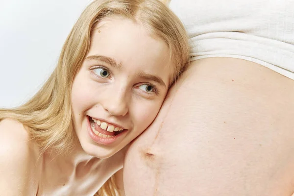 Een blond blank meisje knuffelt en luistert naar de buik van haar zwangere moeder. — Stockfoto