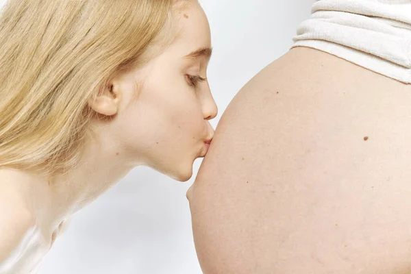 Una ragazza caucasica bionda bacia la pancia della madre incinta isolata. Famiglia e sorella maggiore concetto, vista laterale. — Foto Stock