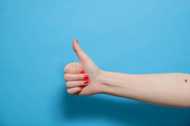 Başparmağını kaldırarak buna benzer bir işaret yapan bir kadın. Mavi arka planda kırmızı jel cilası manikürlü bir kadın eli. Boşluğu kopyala