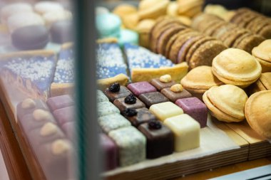 A variety of biscuits, cookies, and petit fours are neatly displayed in a glass bakery showcase. The assortment includes layered pastries, chocolate squares, shortbread. 