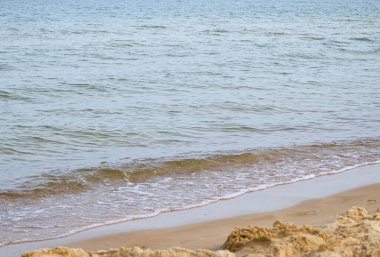 Sakin deniz sahnesi, kumlu bir sahile vuran hafif dalgalar, sahil şeridinde küçük köpük çizgileri ve yumuşak pastel renkler. Huzurlu kıyı manzarası 