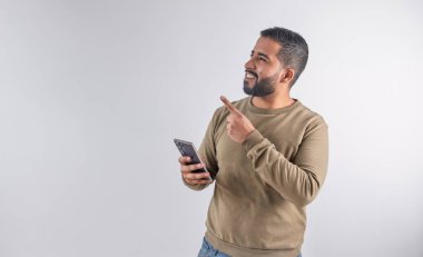 Süveter giyen mutlu bir adam cep telefonunu kullanıyor ve geniş bir kopya alanını işaret ediyor.