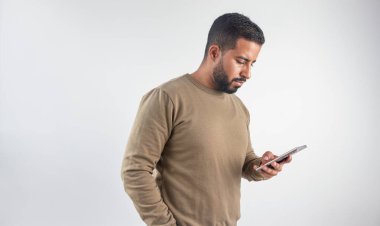 Yoğunlaşmış genç adam aşağıya bakıyor, aktif olarak cep telefonunu temiz beyaz bir arkaplanda kullanıyor.