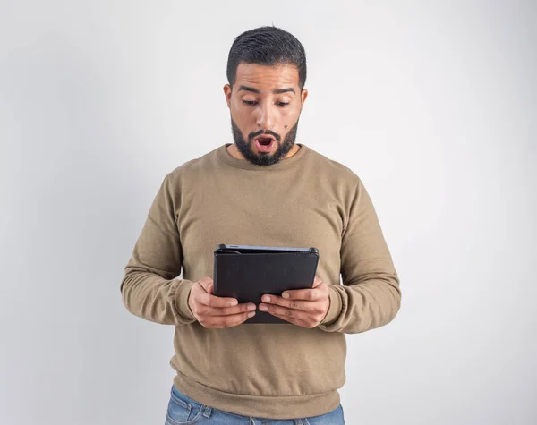 Ağzı açık bir şekilde dijital tabletten beklenmedik haberler izleyen şaşırmış genç bir adamın ön görüntüsü.